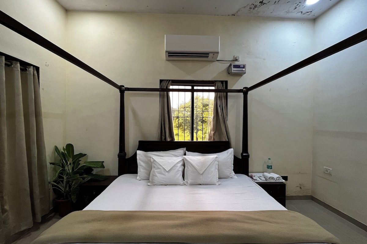 A four-poster bed with white linens, a bedside table with a phone and a bottle, and a window with curtains.