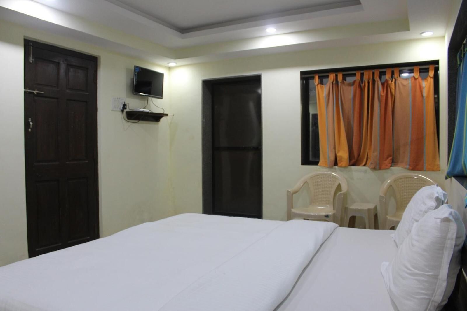 A hotel room with a bed, a TV mounted on the wall, two chairs, and a window with curtains.