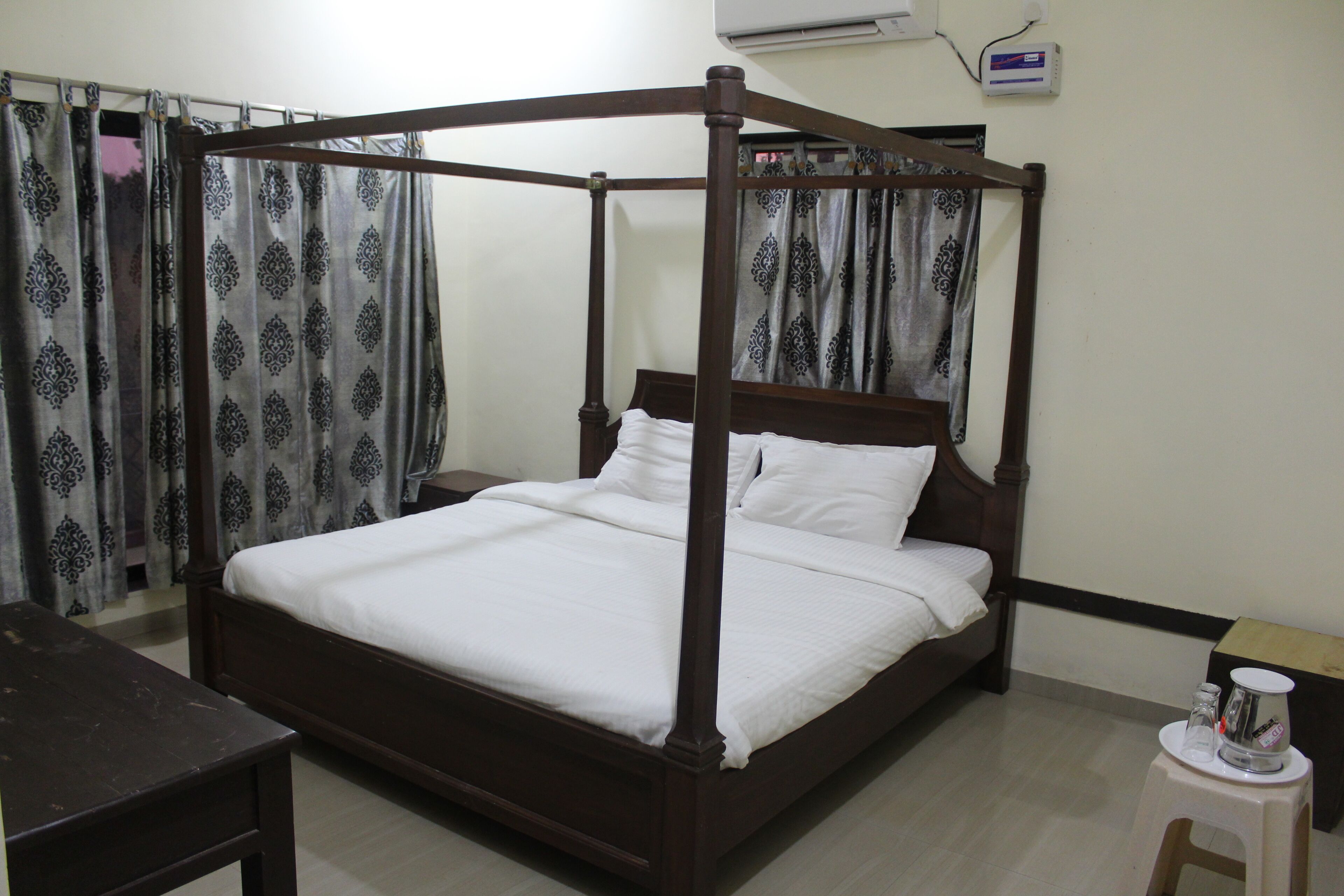 A four-poster bed with white bedding, a wooden nightstand, a small table with a cup and a book, and patterned curtains.