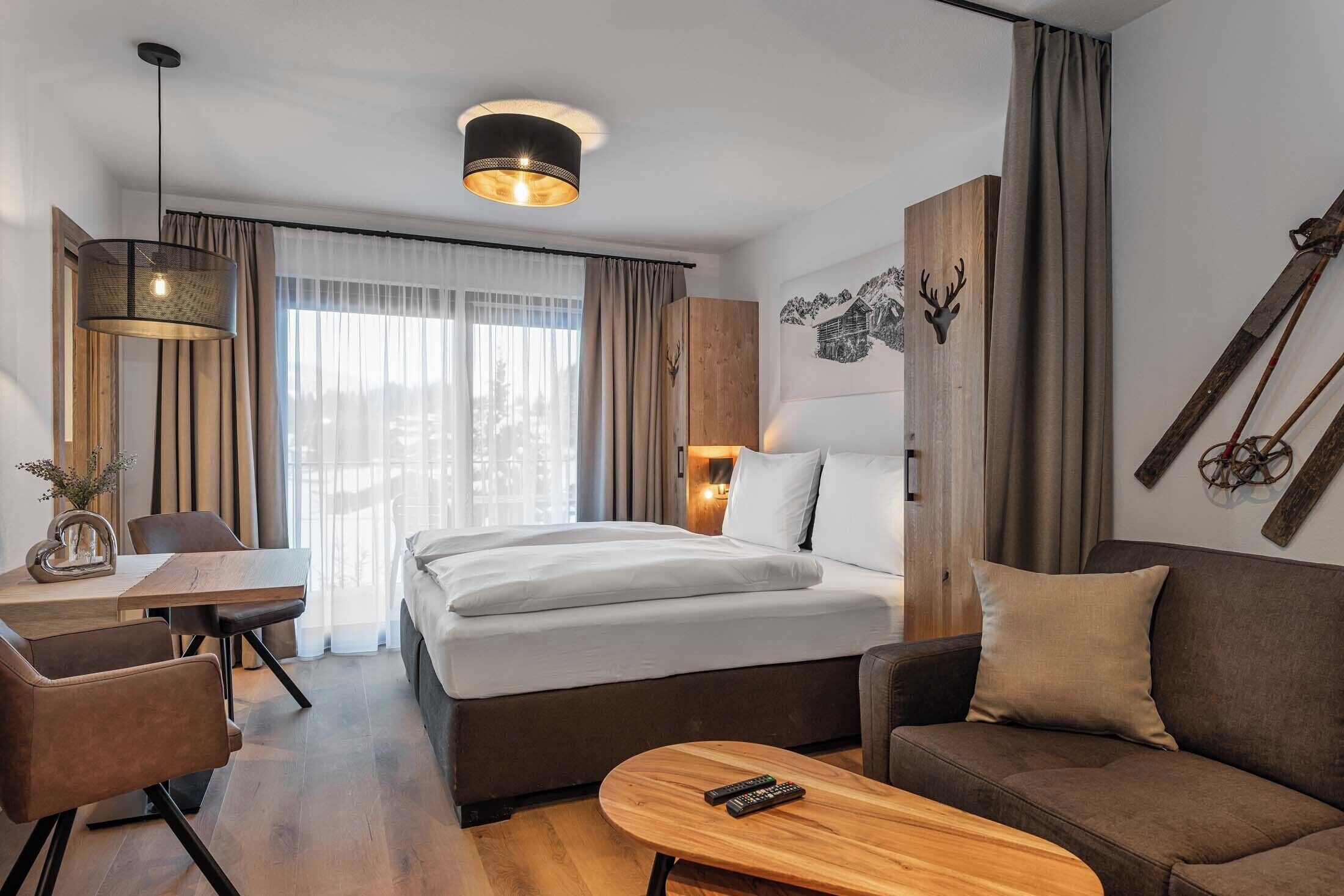 A modern hotel room with a large bed, a sofa, a dining table with chairs, and a small coffee table. There is a window with curtains, a wall-mounted lamp, and a decorative wall piece.