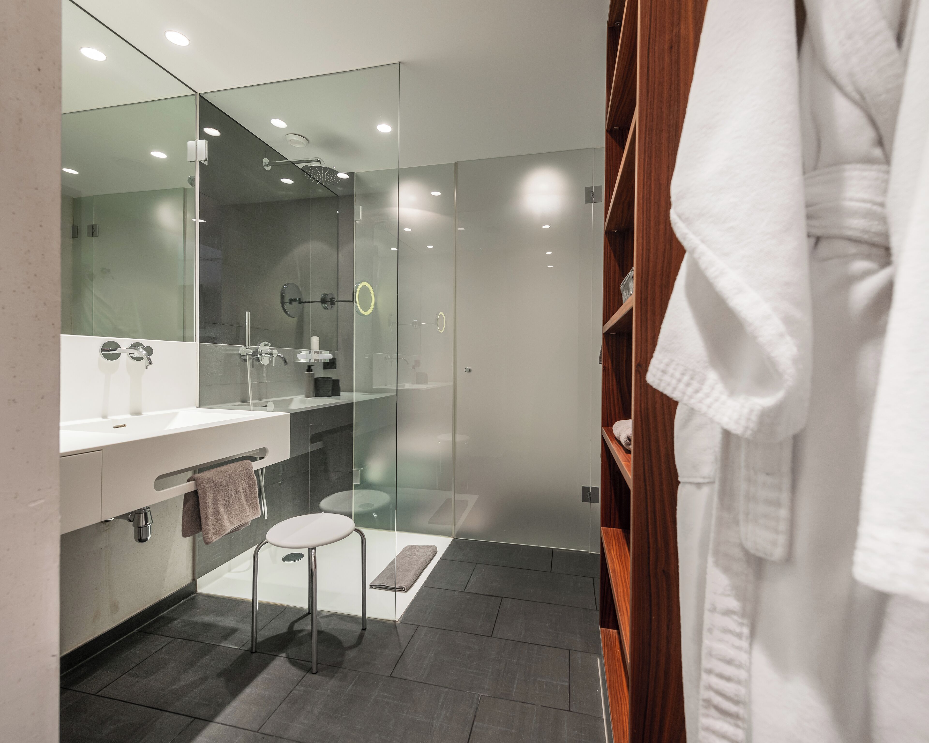 A modern bathroom with a glass-enclosed shower, a vanity with a mirror, and a wooden robe stand.