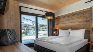 A bedroom with a bed, a wooden headboard, a large window with a view of a snowy landscape, and a hanging light fixture. - AlpenParks Hotel & Apartment Taxacher (Kirchberg in Tirol)