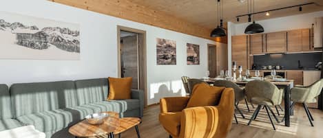 Una sala de estar moderna con un sofá, sillones y una mesa de centro. Hay una zona de cocina con armarios de madera y una mesa de comedor.