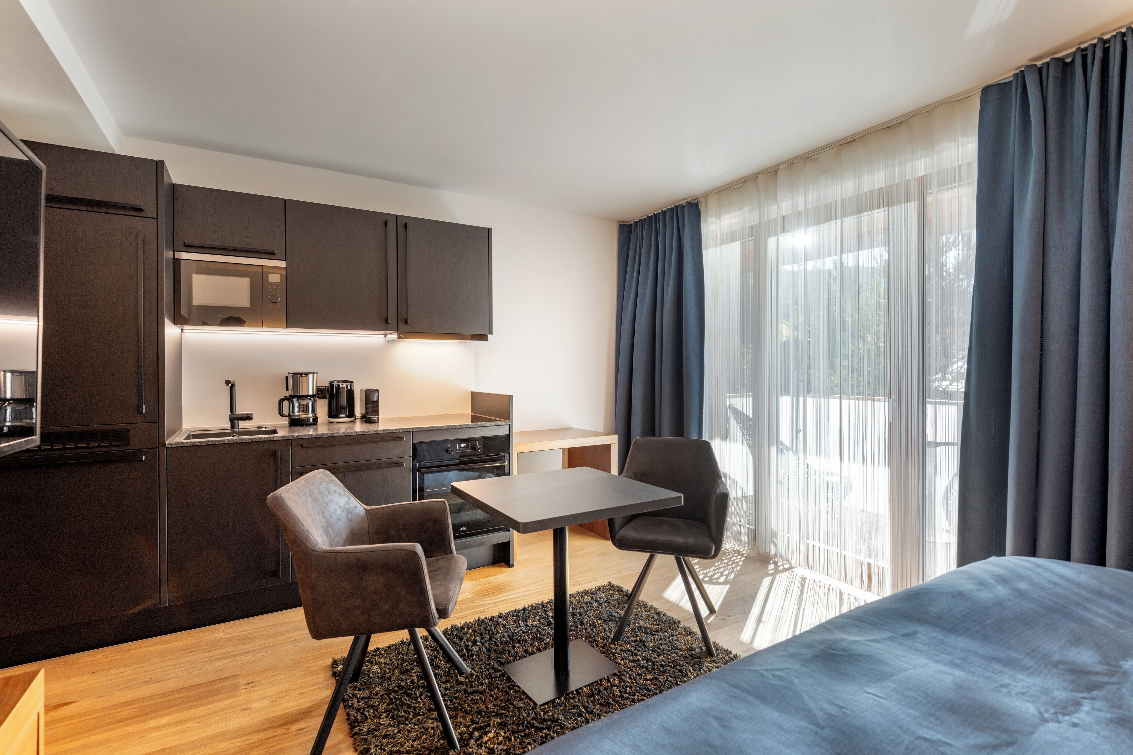 A modern hotel room with a kitchenette, a dining area, and a large window with curtains.