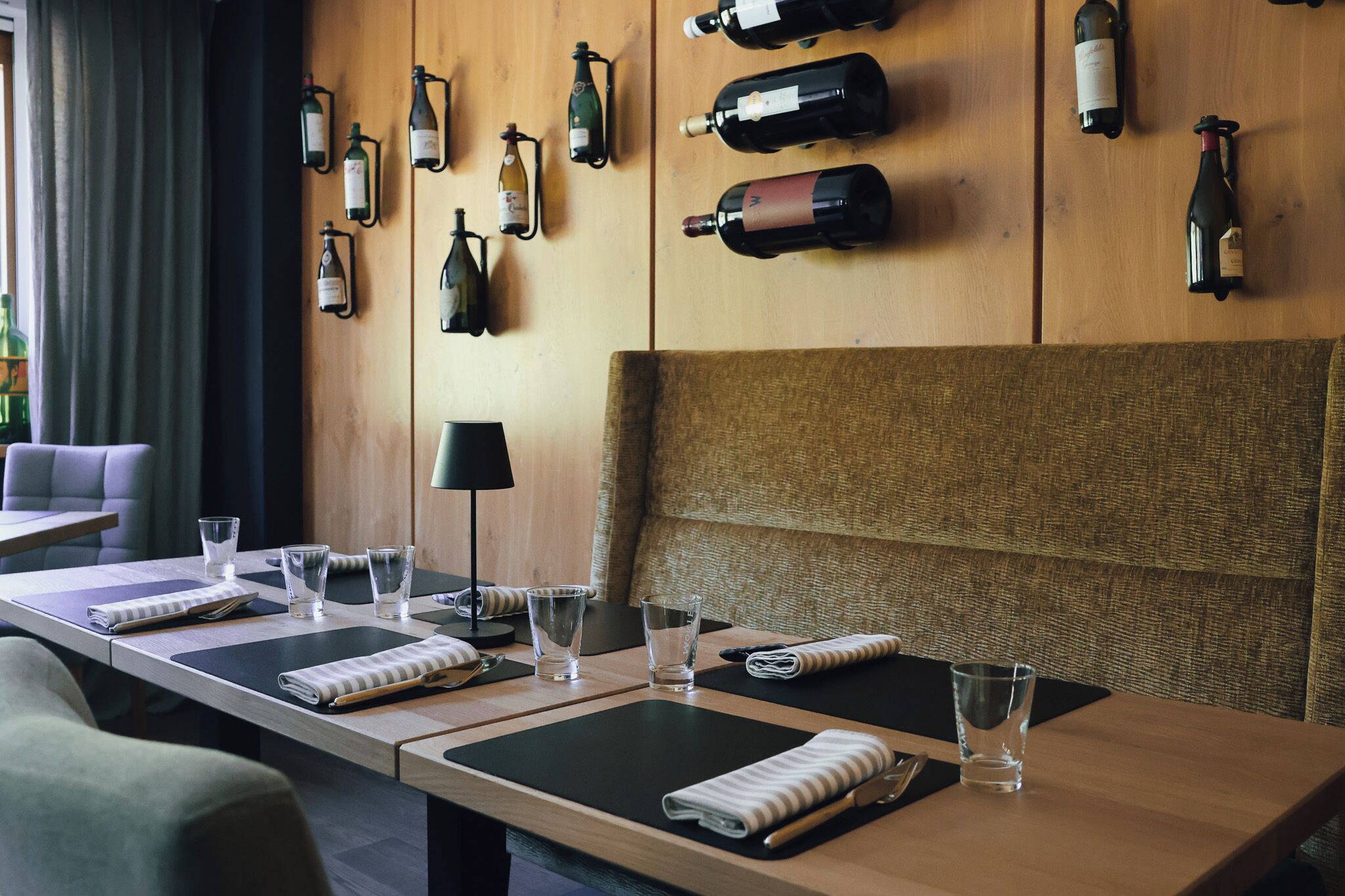 Habitación de hotel con una larga mesa de comedor preparada para dos personas, un sofá y una pared decorada con botellas de vino.