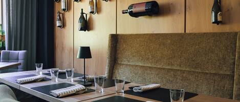 Habitación de hotel con una larga mesa de comedor preparada para dos personas, un sofá y una pared decorada con botellas de vino.