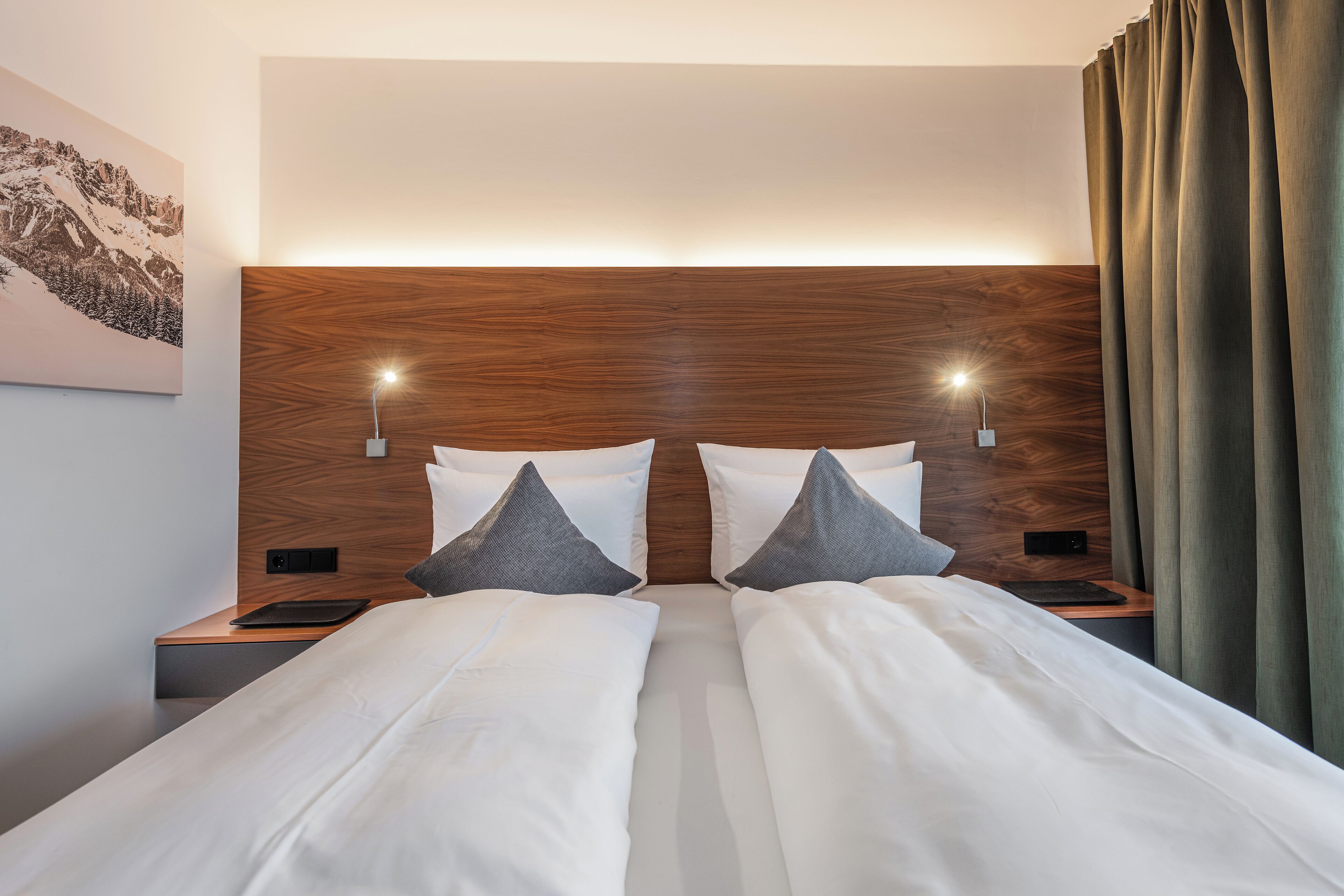 A neatly made bed with white linens and two pillows, a wooden headboard, bedside lamps, and a wall-mounted light.