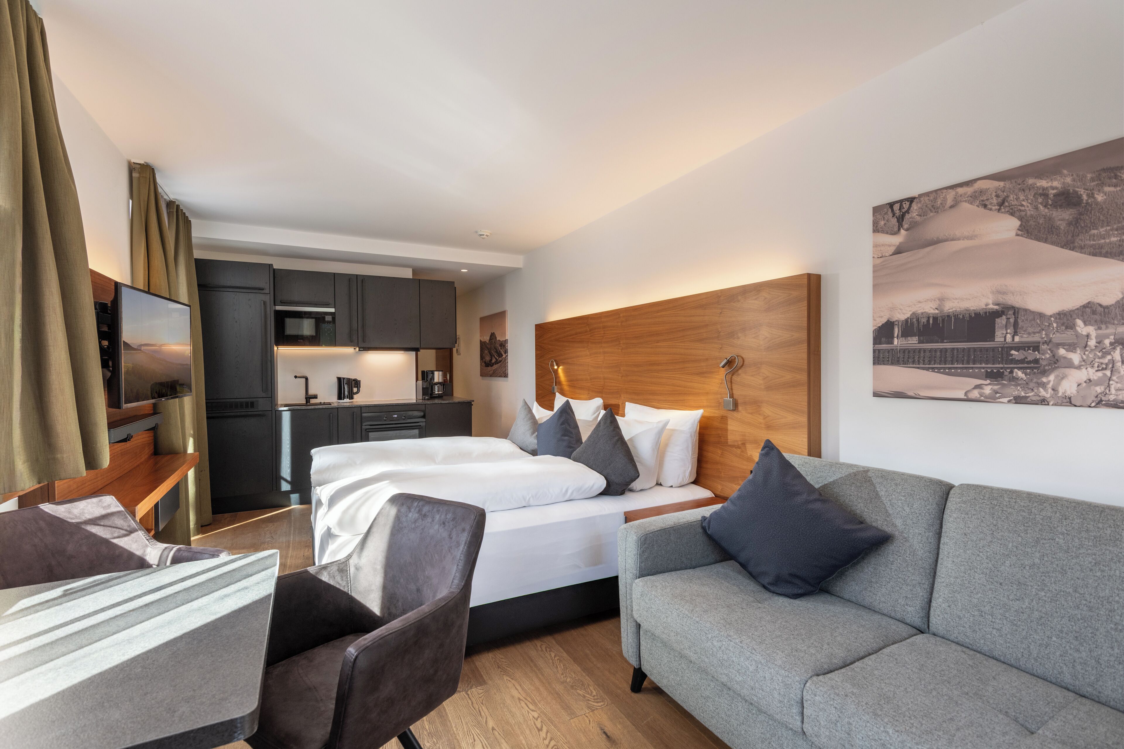 A modern hotel room with a bed, a sofa, a desk, and a kitchen area.