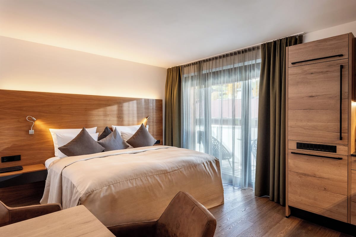 A hotel room with a large bed, wooden headboard, a desk with a chair, a wardrobe, and a balcony with curtains.