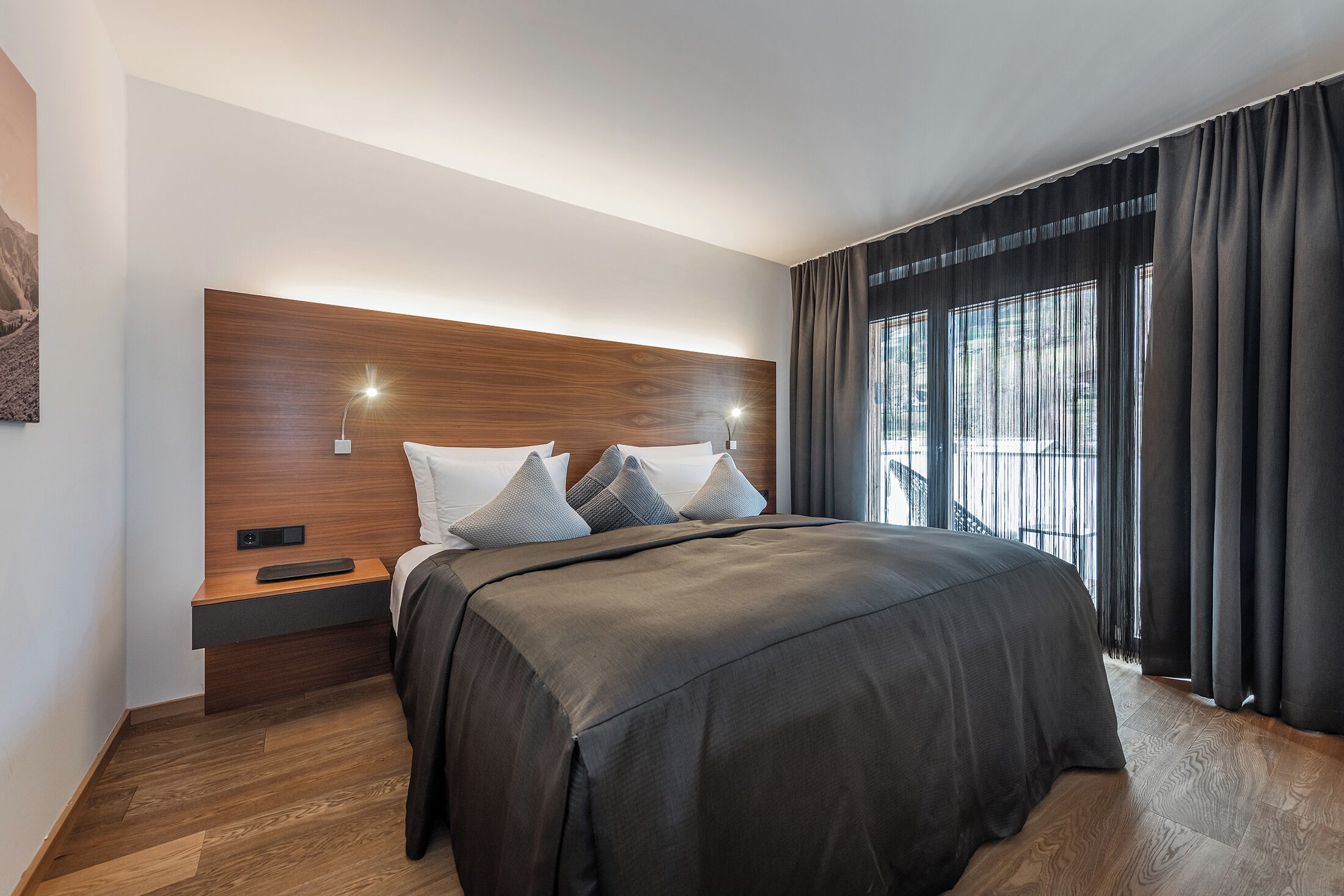 A modern bedroom with a large bed, a wooden headboard, bedside tables, and a view of the outdoors through large windows.