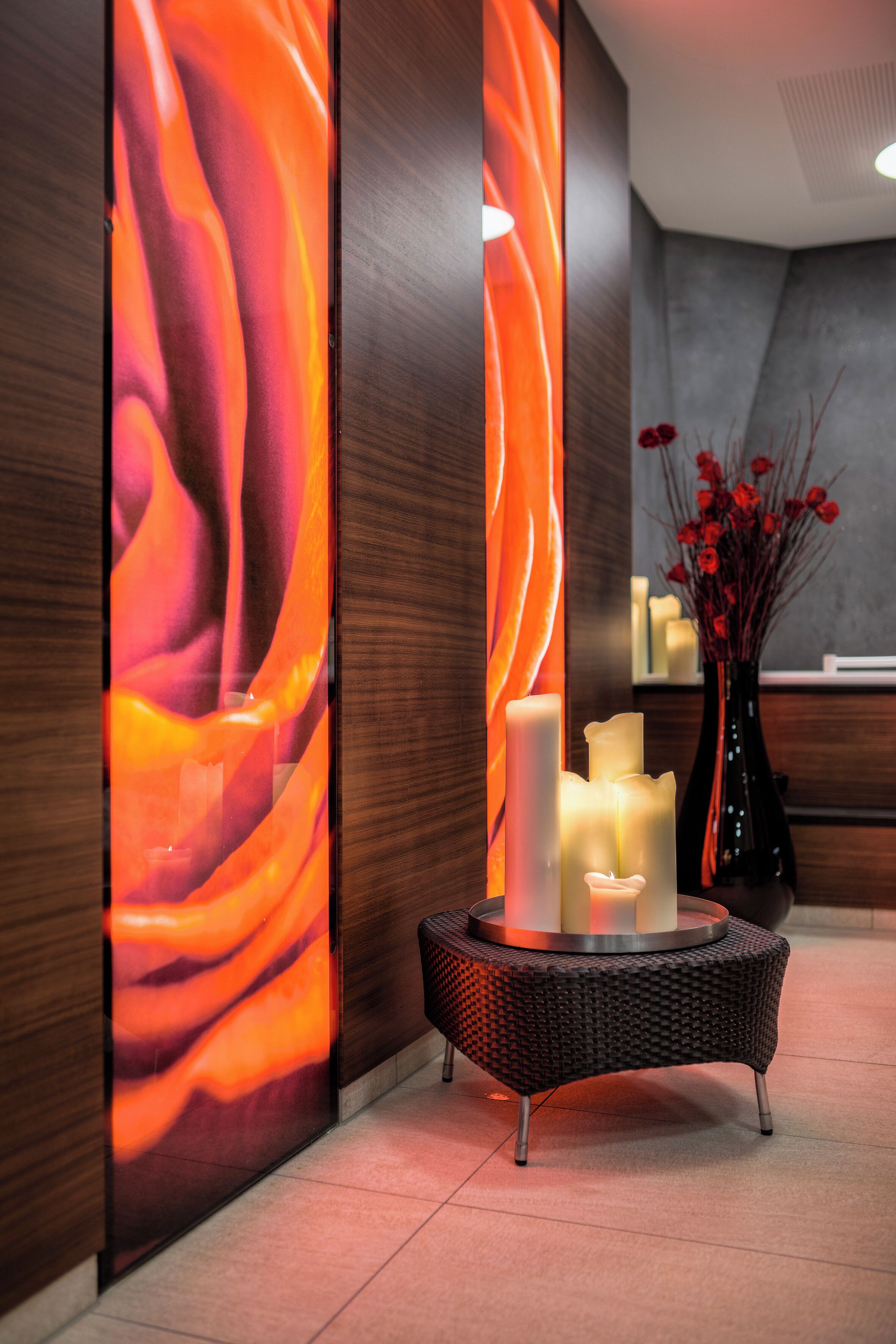 A reception area with a modern design, featuring a wall with a dynamic abstract pattern, a round wicker table with candles, and a vase with red flowers.