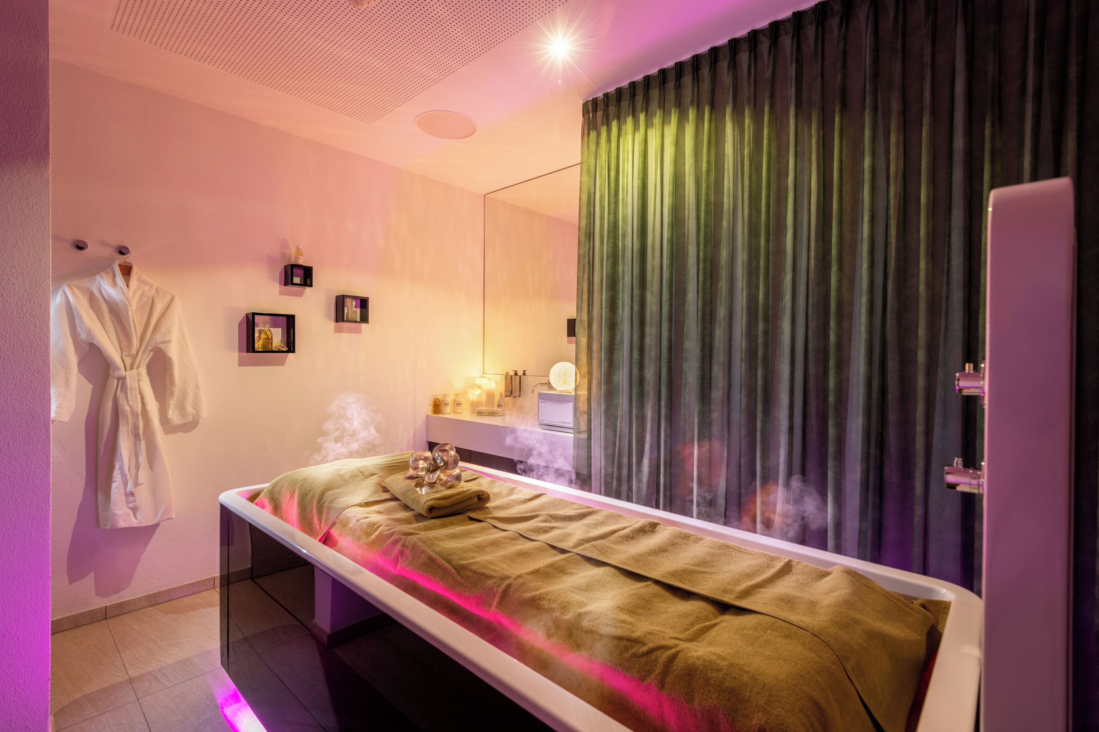 A modern spa room with a massage table, a robe hanging on the wall, and a mirror reflecting the room.