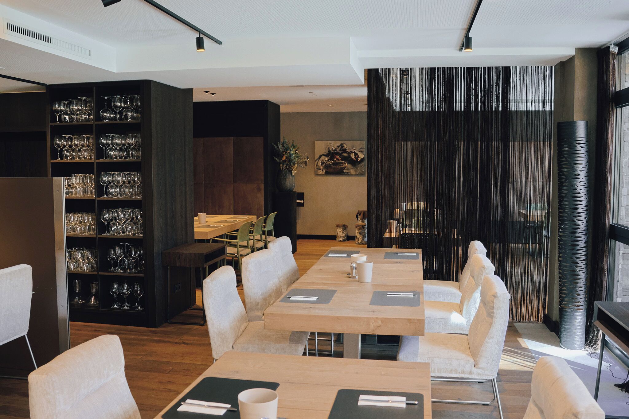 A modern dining area with wooden tables and white chairs, a bookshelf with glassware, and a decorative wall with vertical lines.