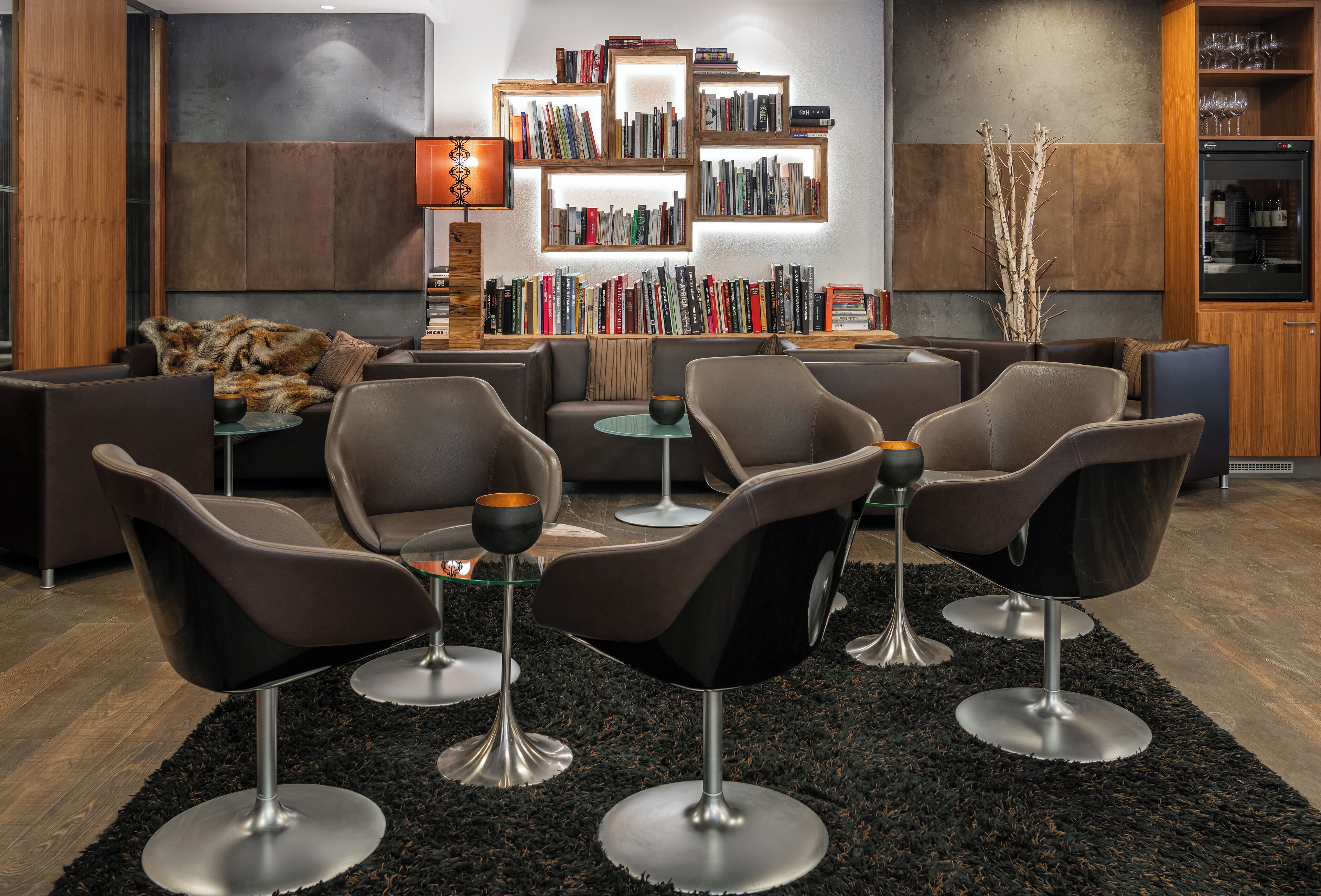 Een moderne zithoek met leren banken, metalen stoelen, een centrale salontafel en een boekenkast vol boeken.