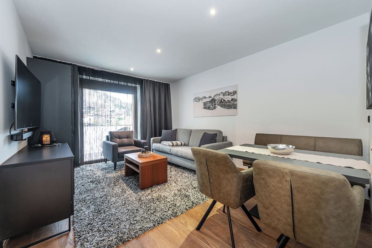 A modern hotel room with a sofa, a dining table, a TV, and a large window with curtains.