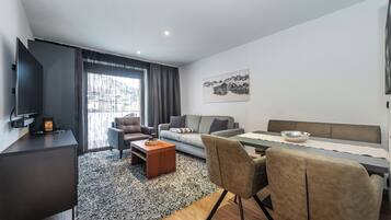 Una habitación de hotel moderna con sofá, mesa de comedor, televisor y un amplio ventanal con cortinas.