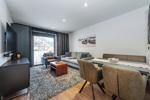 Una habitación de hotel moderna con sofá, mesa de comedor, televisor y un amplio ventanal con cortinas.