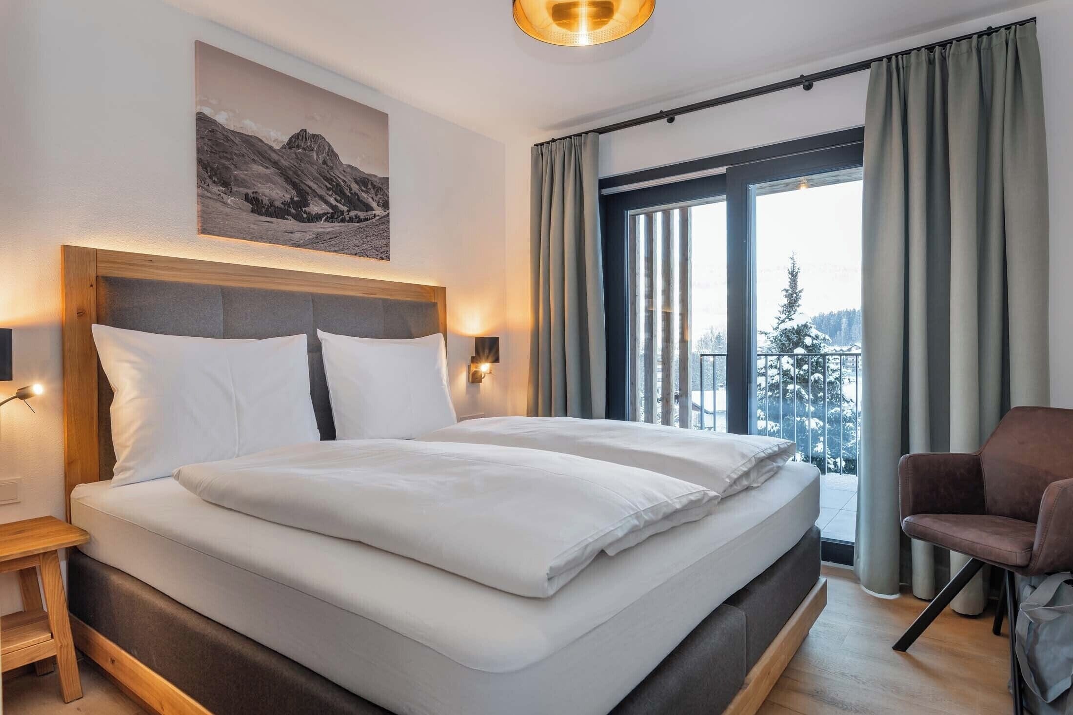 A modern hotel room with a large bed, a nightstand, a chair, and a window with curtains.