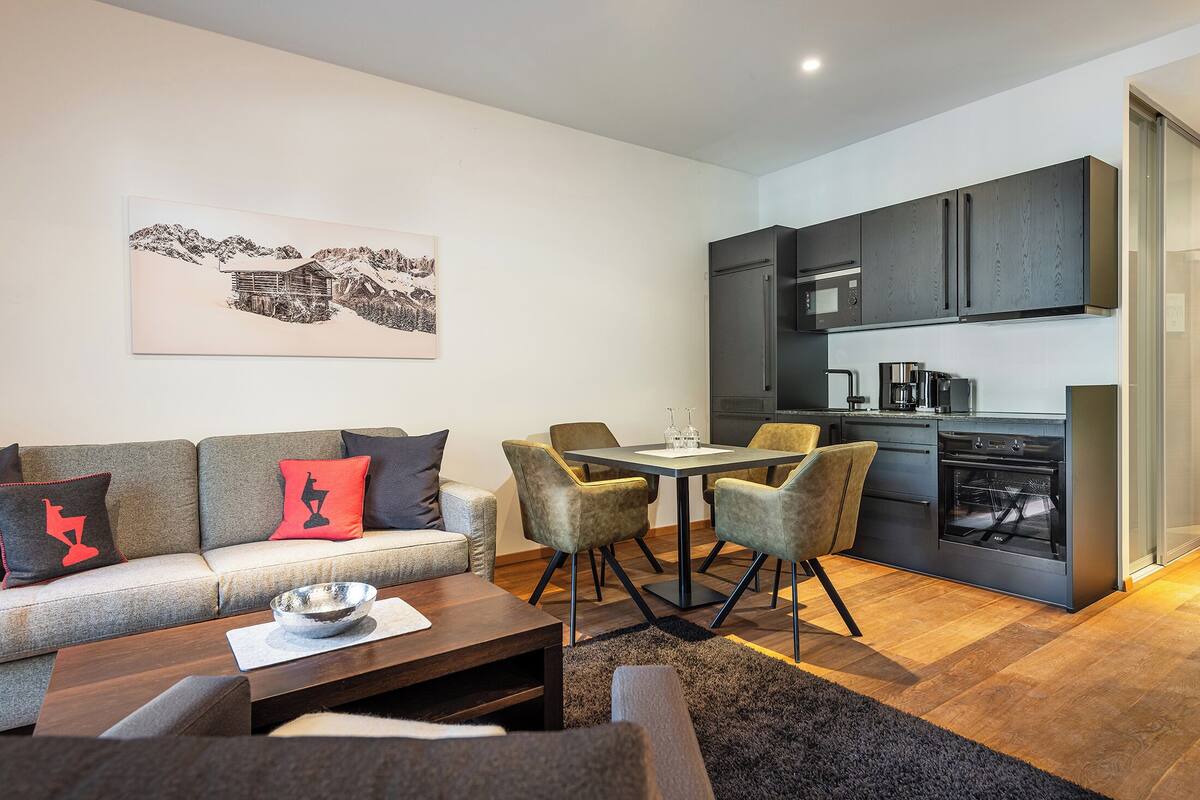 A modern living room with a sofa, coffee table, and chairs, a dining table with chairs, and a kitchen area with cabinets and appliances.