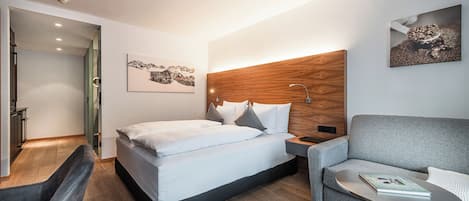 Quarto de hotel moderno com uma cama, um sofá cinzento, uma mesinha e uma cabeceira de madeira.