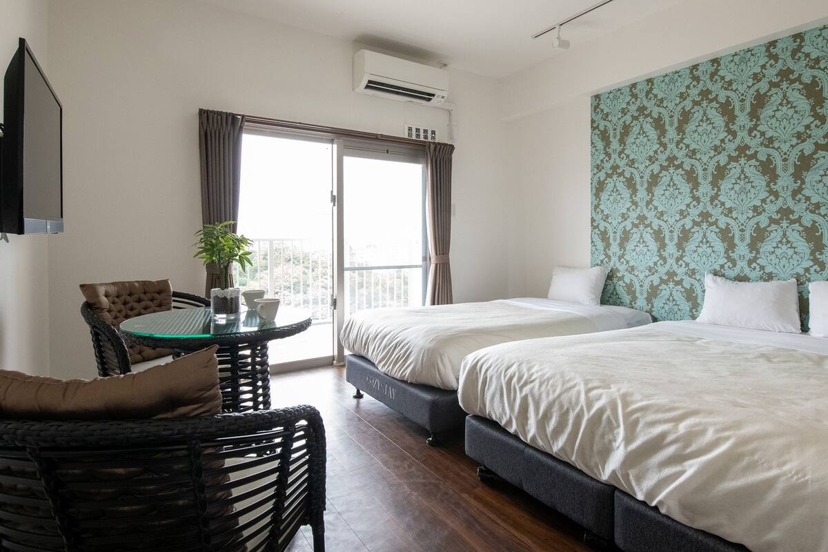 A hotel room with two beds, a balcony with curtains, a small table with a plant, and a TV mounted on the wall.
