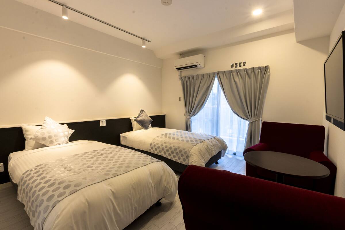 A hotel room with two beds, a red chair, a small table, and a large window with curtains.