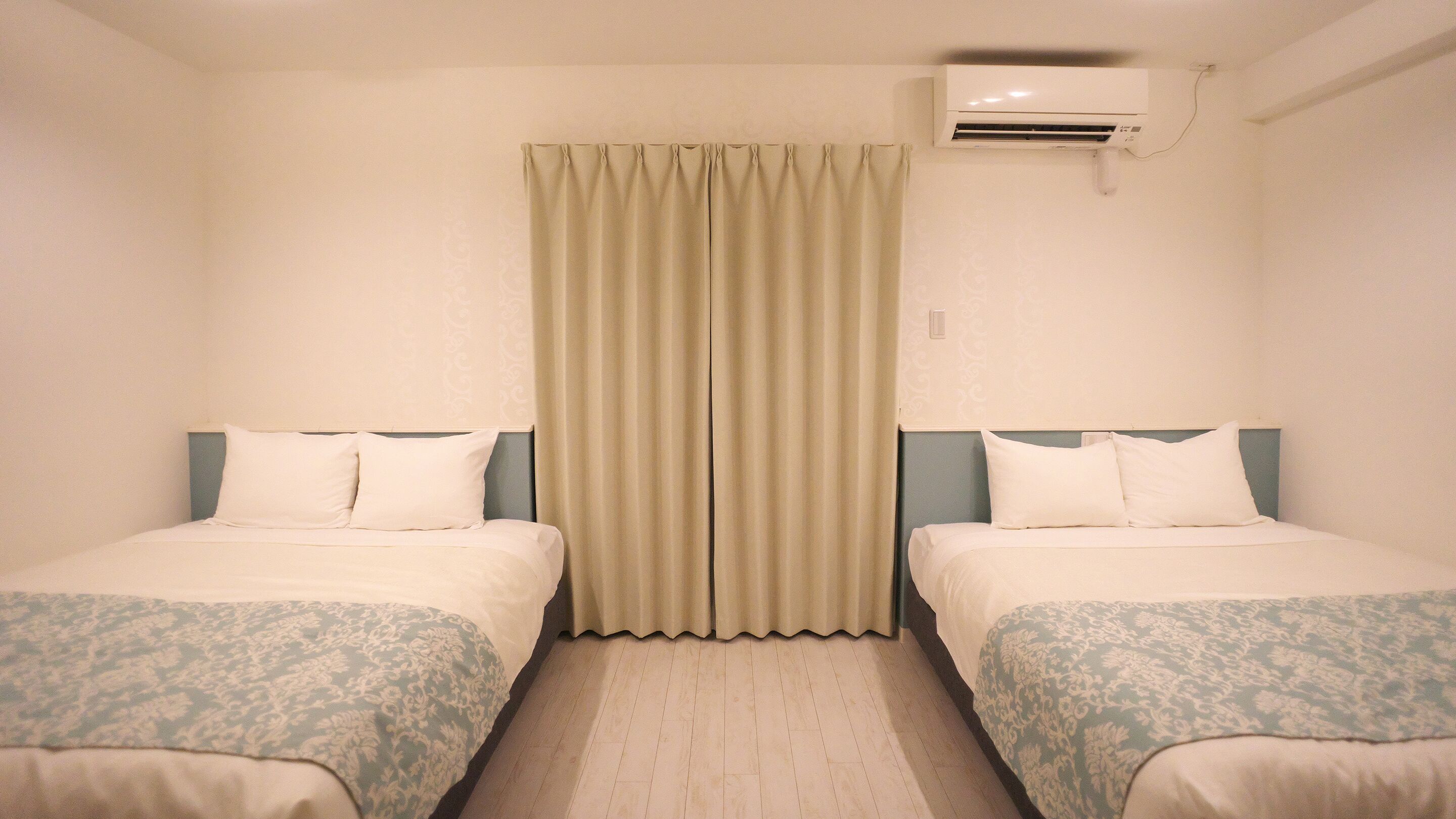 A hotel room with two beds, a curtain, an air conditioning unit, and a patterned bedspread.