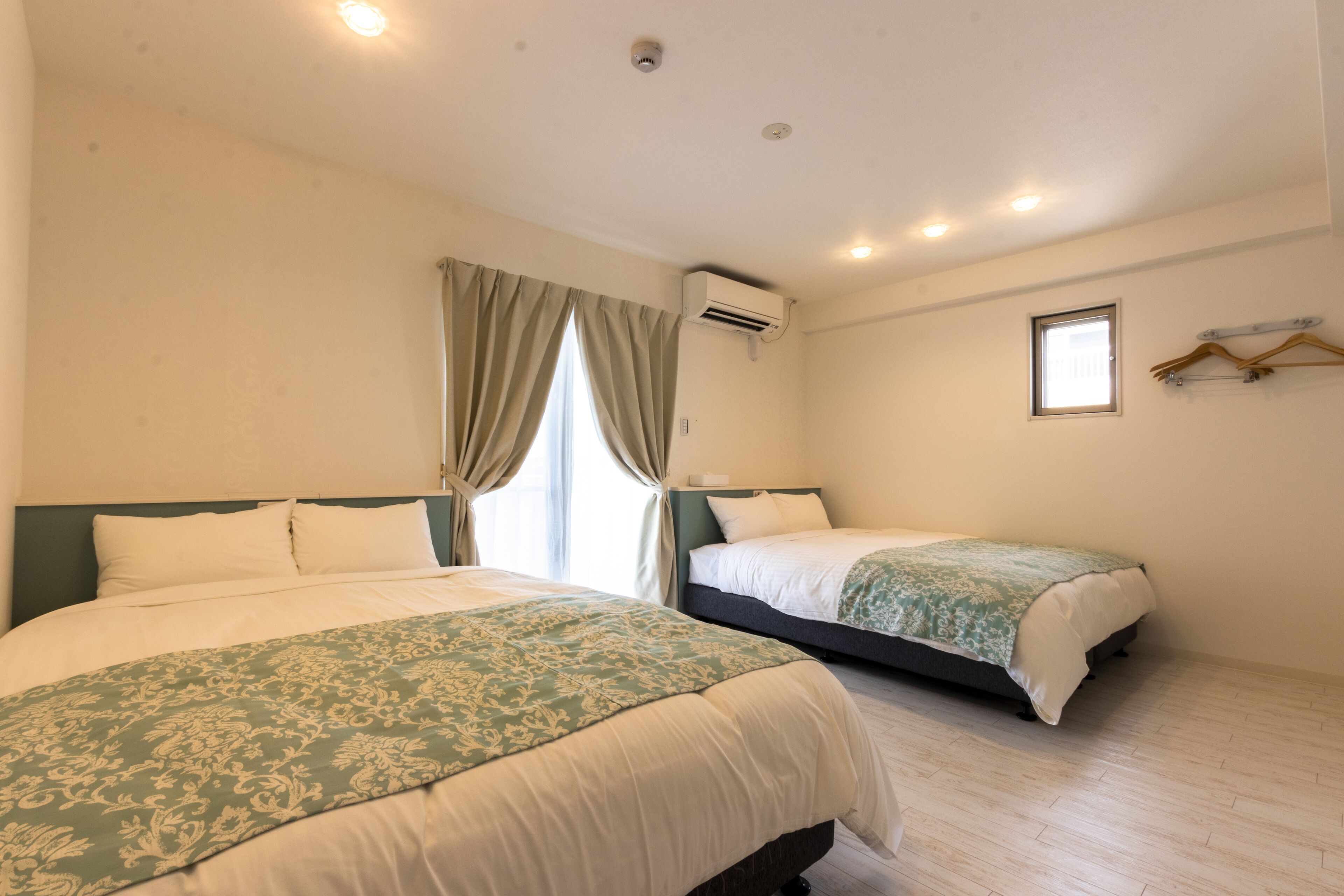 A hotel room with two beds, a window with curtains, an air conditioning unit, and a wall-mounted hanger.