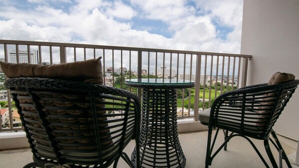 A balcony with two wicker chairs and a small round table, overlooking a cityscape with buildings and greenery.