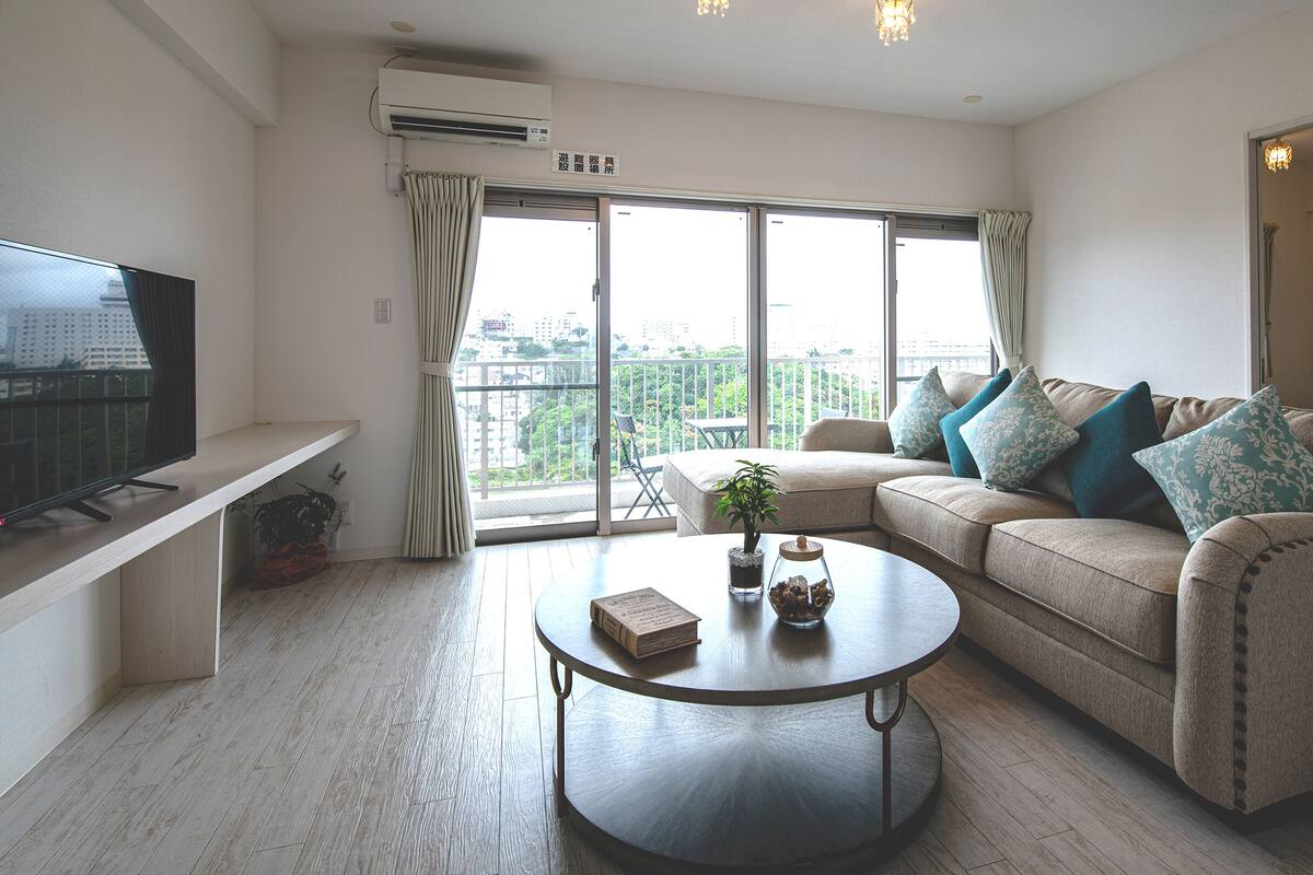 A modern living room with a sofa, a coffee table, a TV, and a balcony with a view of the city.