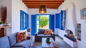 Un salon chaleureux avec un canapé, un fauteuil et une table basse, des poutres en bois au plafond et une vue à travers des volets bleus.