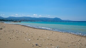 Une plage à l’eau d’un bleu limpide, bordée de galets, avec des montagnes qui se dessinent à l’horizon.
