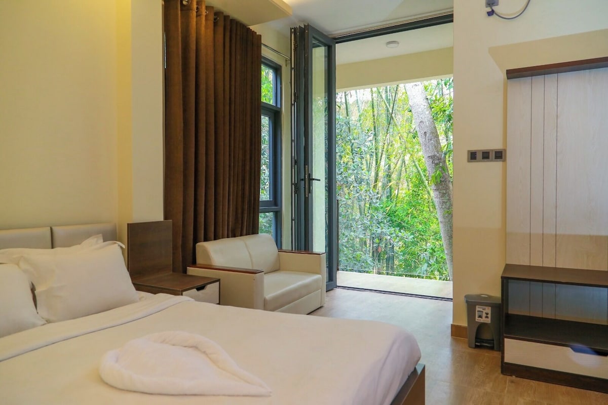 A modern hotel room with a large bed, a seating area, and a balcony with a view of greenery.