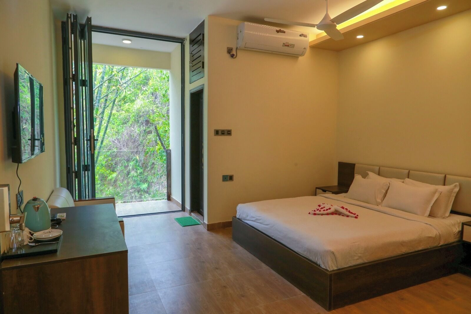 A bedroom with a bed, a desk, a chair, an air conditioning unit, and a view of greenery outside.