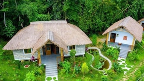 Two thatched-roof cottages surrounded by lush greenery, with a garden path leading to each house. - Balishira Resort (Kamalganj)