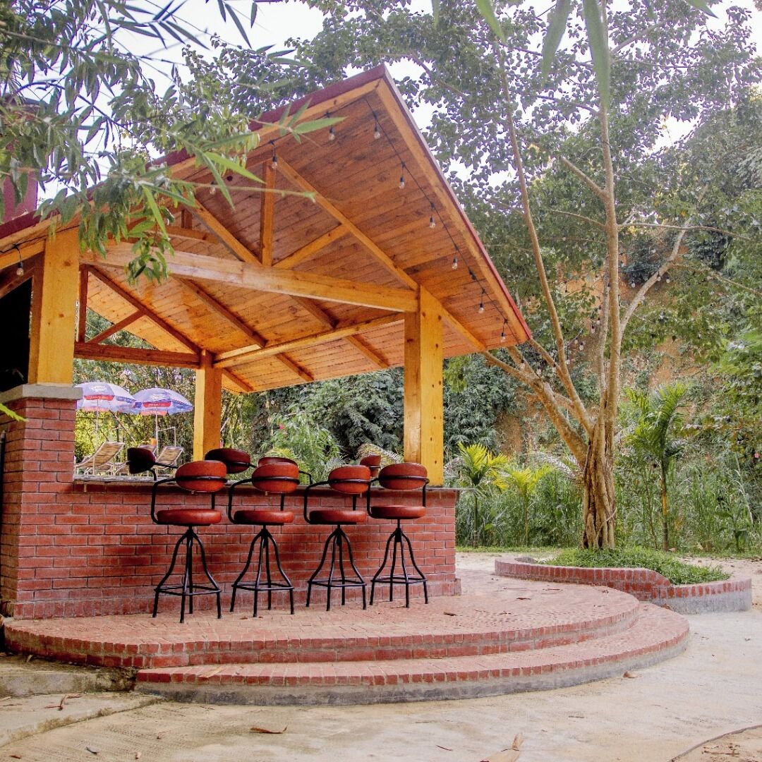 An outdoor barbecue area with a brick counter and stools, surrounded by greenery.