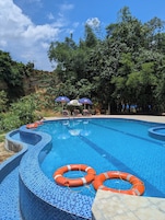 Piscina com azulejos de mosaico azul, rodeada por árvores e um céu claro.