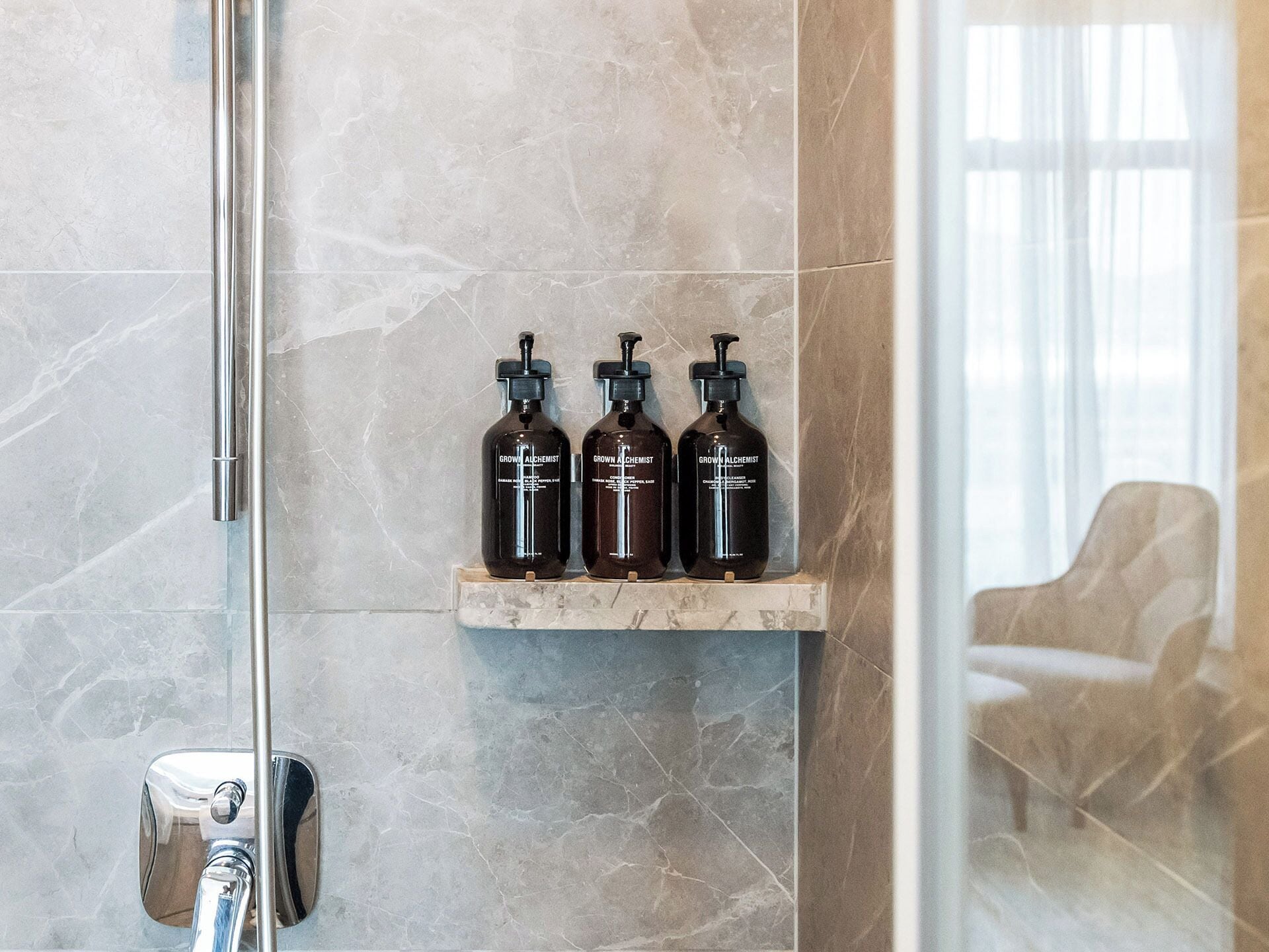 Un baño con pared de mármol, cabina de ducha con mampara de vidrio y un estante con tres productos de 'Grown Alchemist'.