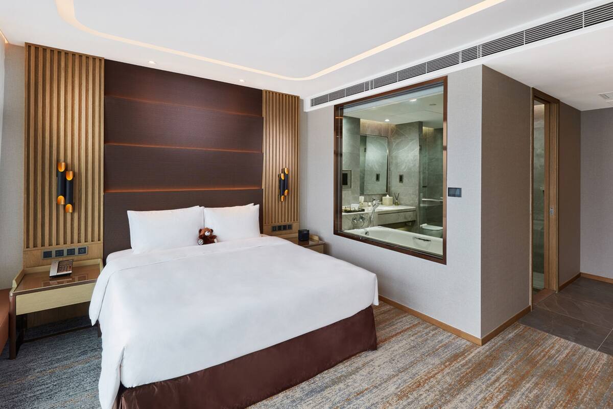 A modern hotel room with a large bed, a nightstand, a desk, and a large mirror reflecting a bathroom.