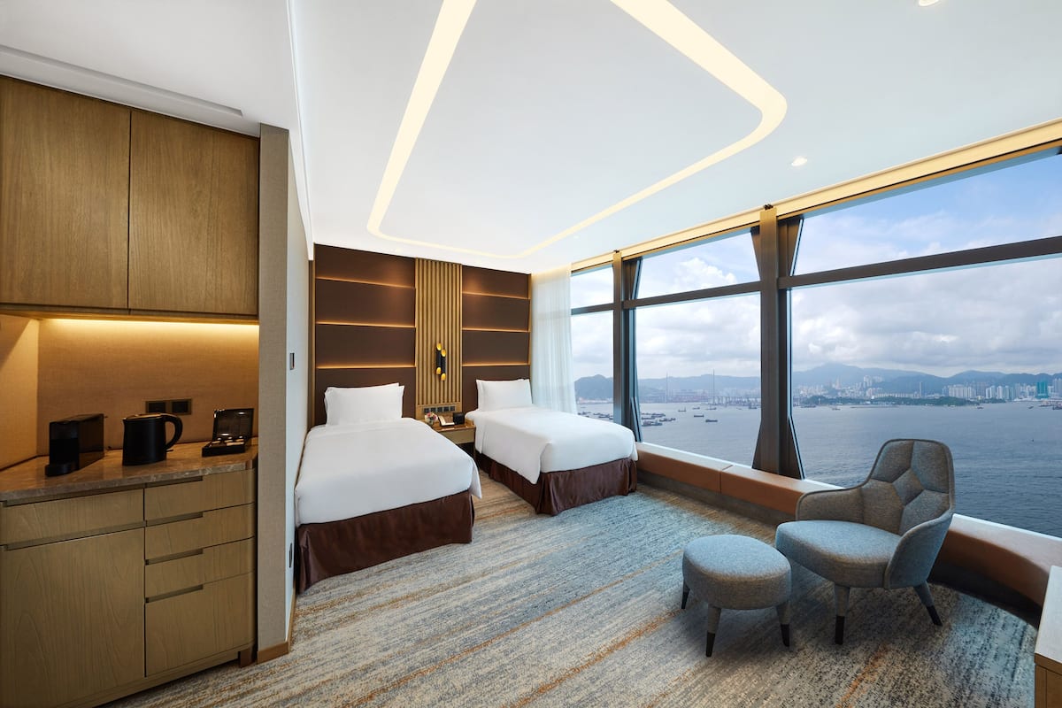 A modern hotel room with two beds, a desk, a chair, and a large window offering a city view.