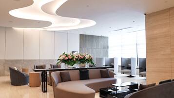 A modern lobby with a curved ceiling, a seating area with sofas and chairs, a reception desk, and a large window providing natural light.
