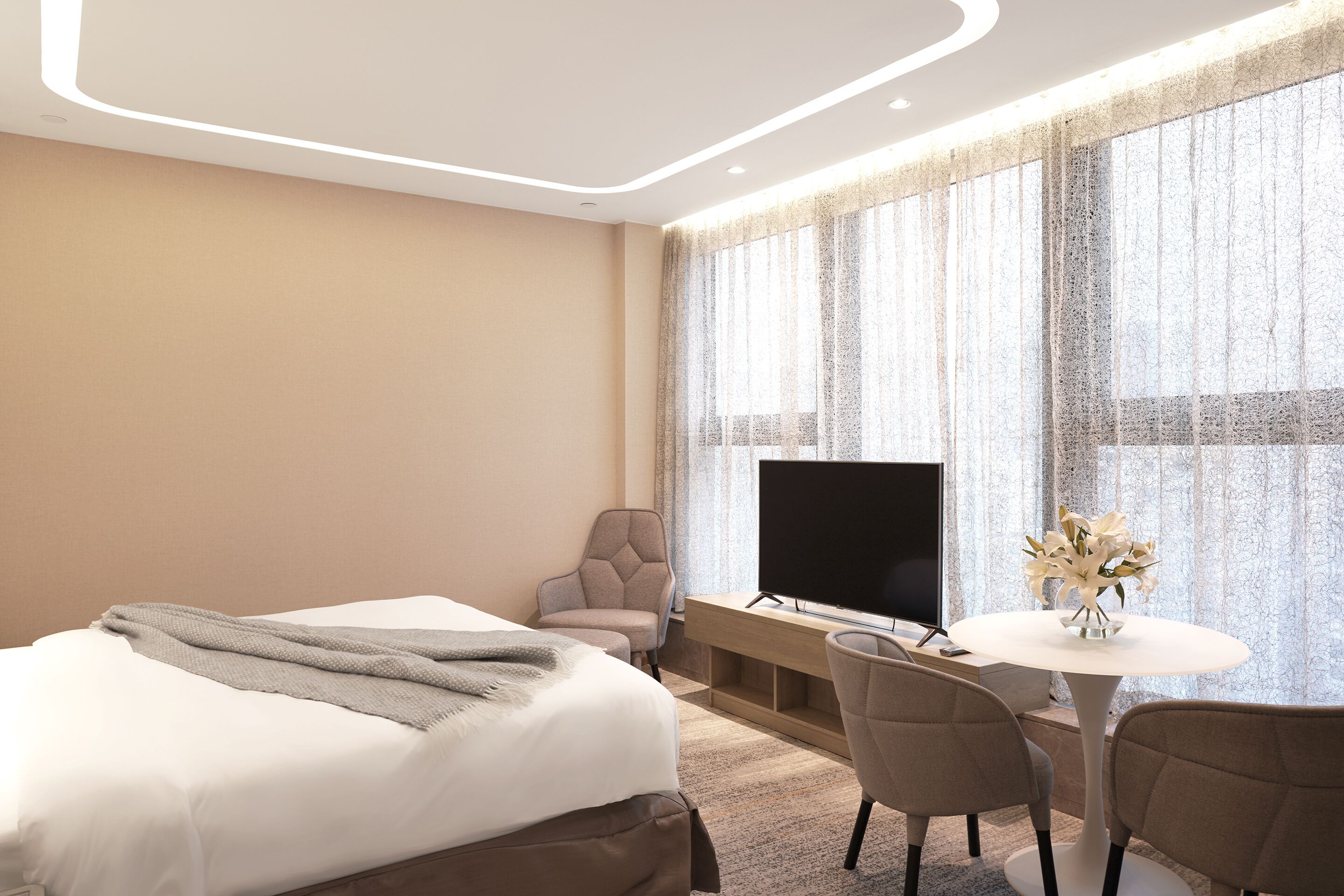 A modern hotel room with a large bed, a desk with a flat-screen TV, a seating area with chairs, and a large window with sheer curtains.