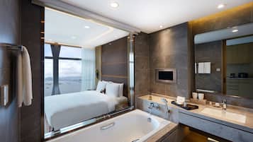 A modern hotel bathroom with a large bathtub, a vanity with a mirror, and a view of a bed with a cityscape.