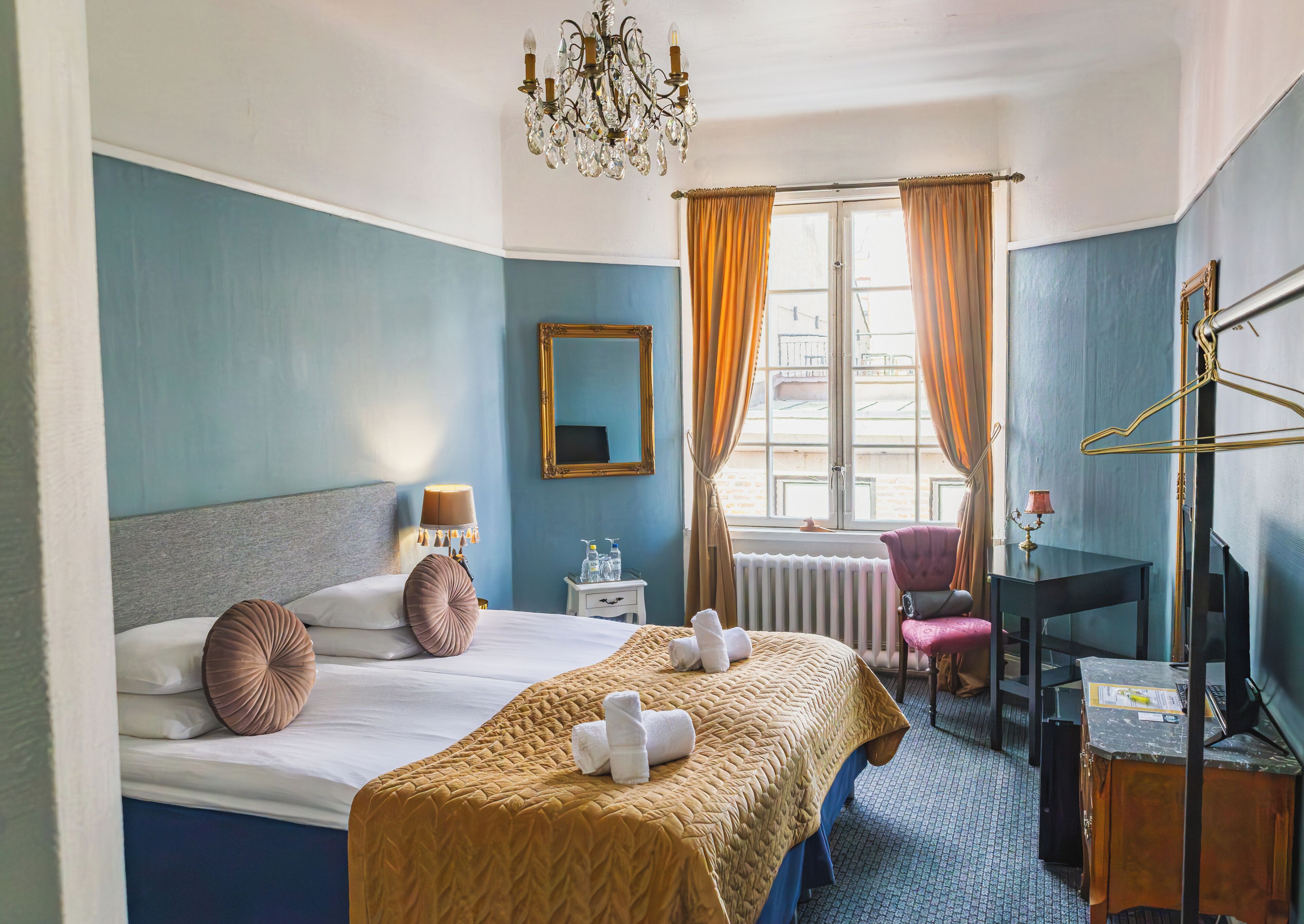 Habitación de hotel con cama, escritorio, silla, espejo, radiador y ventana con cortinas.