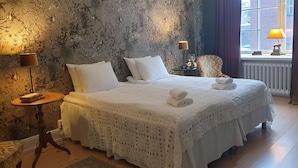 Ein ordentlich bezogenes Bett mit weißen Leinentüchern und einer Spitzendecke, ein kleiner runder Tisch mit einer Lampe sowie ein Stuhl mit gemustertem Bezug.