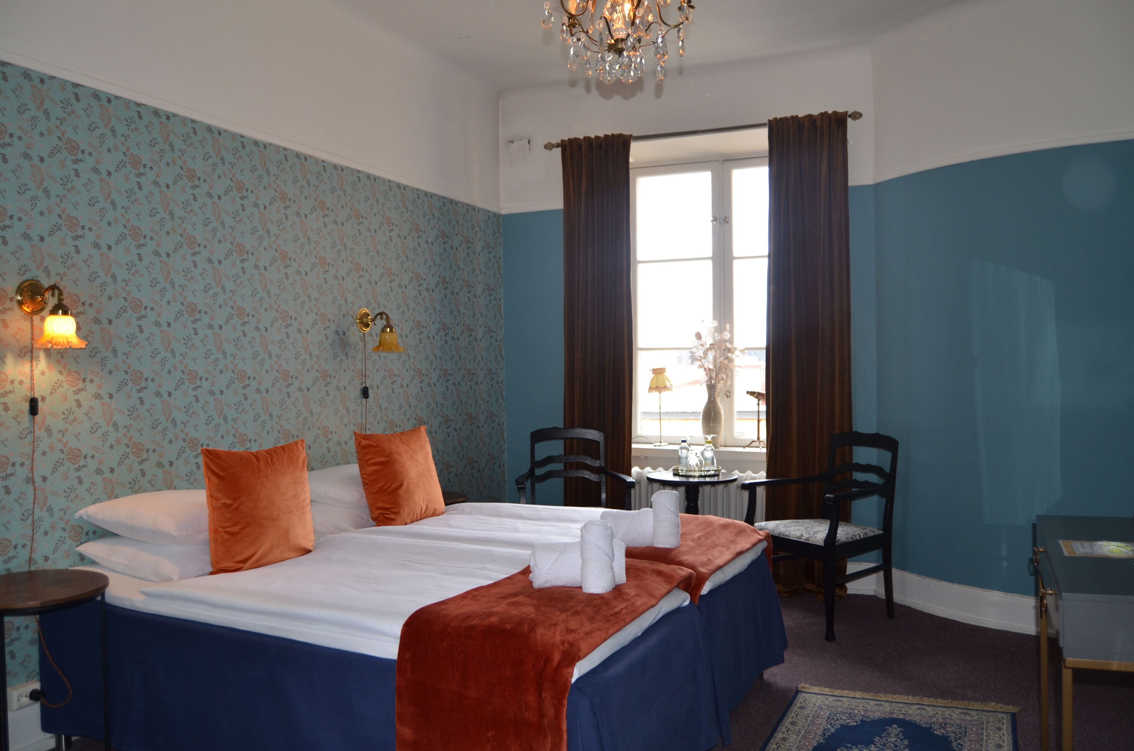 Habitación de hotel con cama doble, dos sillones, una mesita, candelabro y ventana con cortinas.
