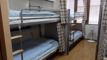 A dormitory room with bunk beds, a standing fan, and a window with blinds.