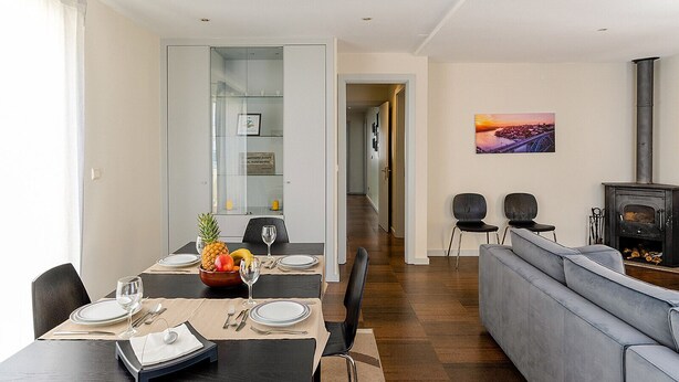 Un salon moderne avec un canapé gris, un coin repas avec une table dressée pour deux personnes et un poêle à bois.