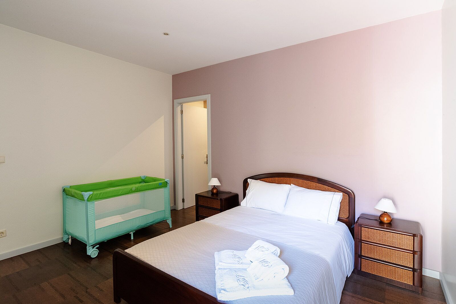 Quarto com cama, berço verde para bebê, duas mesas de cabeceira com abajures e uma porta.