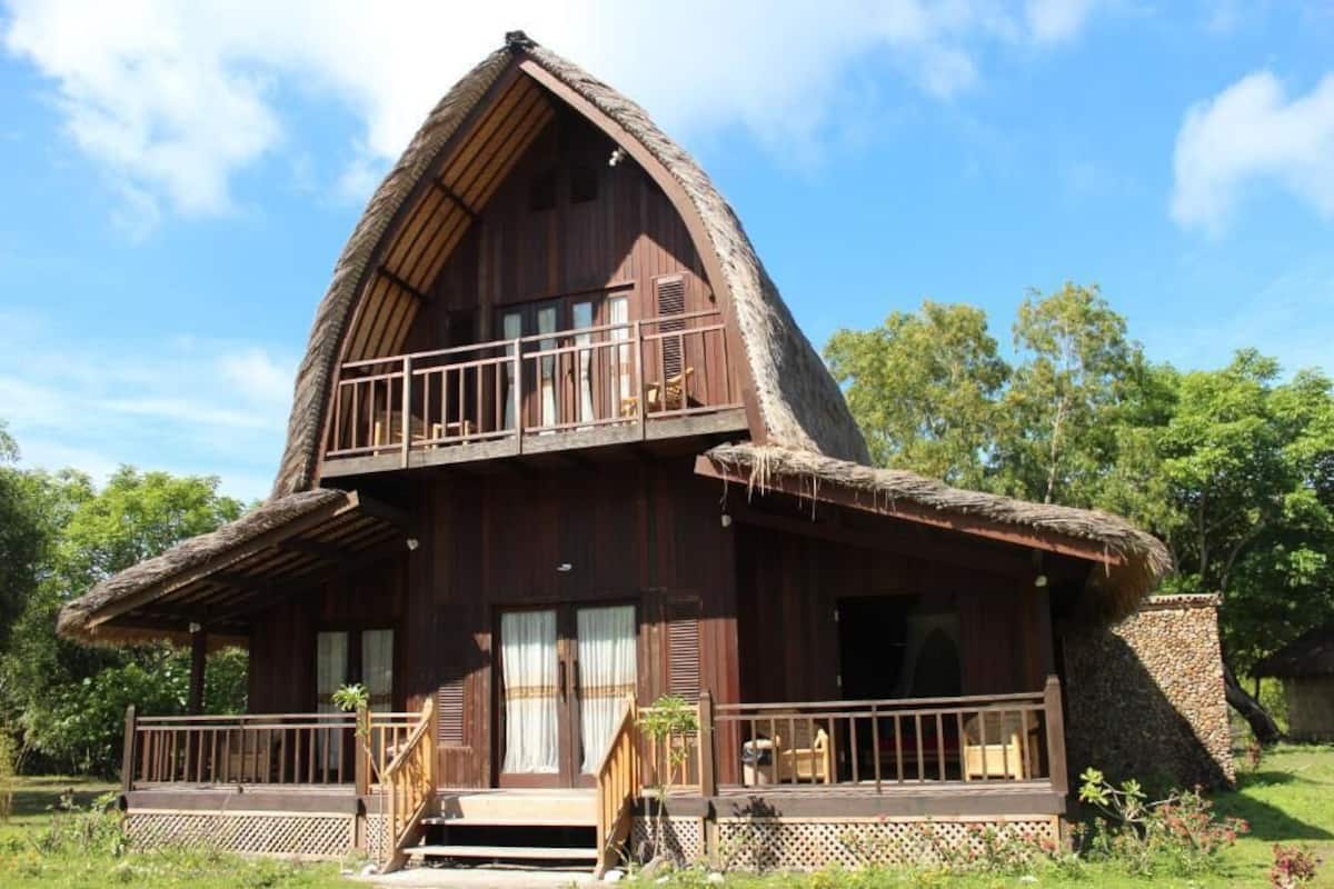 A two-story wooden house with a thatched roof, a balcony on the upper floor, and a front porch with steps.